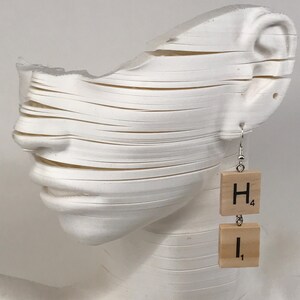 Puede incluir: Pendientes únicos con fichas de Scrabble de madera que deletrean "HI" con valores de puntos. Los pendientes cuelgan de ganchos plateados. Se muestran en una escultura blanca en capas de una cara.