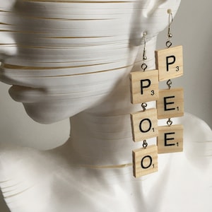 Könnte beinhalten: Ein Paar Hängeohrringe aus hölzernen Scrabble-Kacheln, die das Wort "POOE" buchstabieren. Jede Kachel hat den Buchstaben und den Punktwert aufgedruckt.