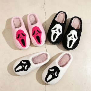 May include: Three pairs of plush, fuzzy slippers in pink, black, and white. Each slipper features a prominent, stylized ghost-like face graphic on the top. The pink pair has a dark pink face, the black pair has a white face, and the white pair has a black face. The interior of each slipper is lined with a soft, light-colored material.
