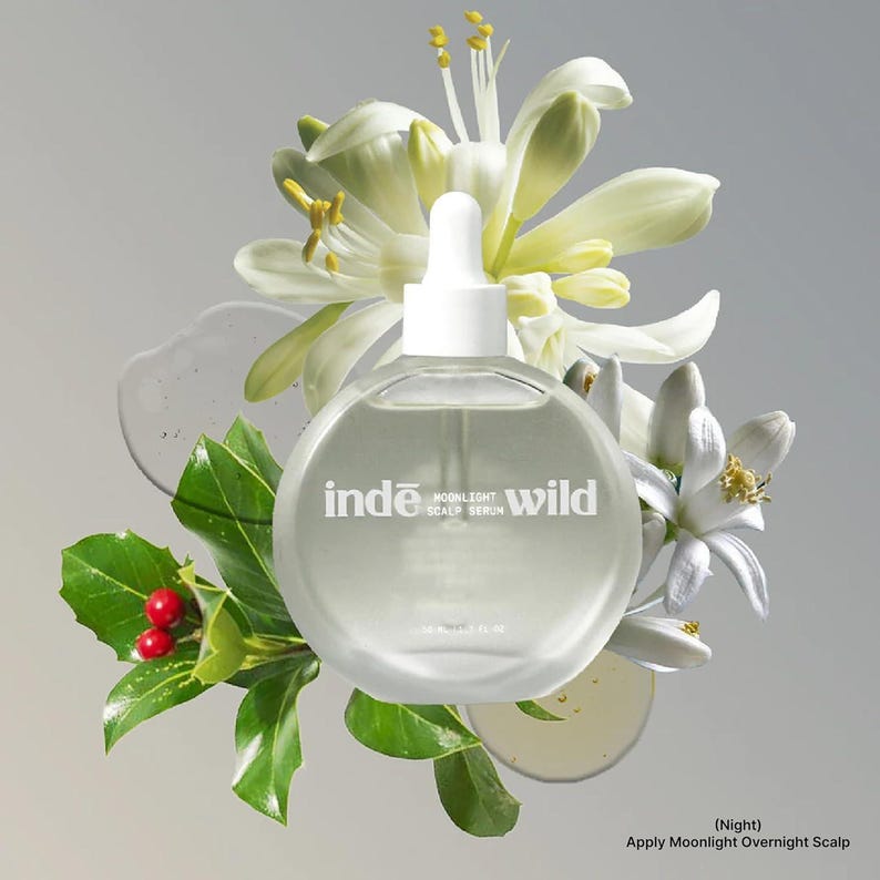 Op de afbeelding: Een heldere, ronde fles Inde Wild Moonlight Scalp Serum met een witte druppelaar, omringd door witte bloemen, groene bladeren en rode bessen. De fles heeft witte tekst met de tekst "Inde Moonlight Scalp Serum Wild".