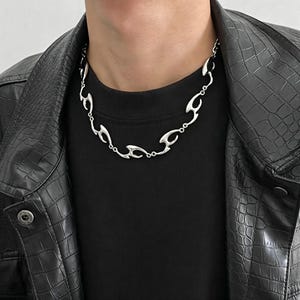 May include: A silver-colored necklace with an abstract flame-like design. The necklace is worn against a black shirt and a black leather jacket. The necklace has a series of interconnected, stylized flame shapes.