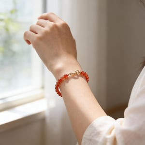 May include: A beaded bracelet featuring small, round, orange-red gemstones. The bracelet is secured with a gold-colored clasp. The image shows the bracelet on a person's wrist, with a soft, natural light illuminating the piece.