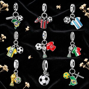 Könnte beinhalten: Eine Sammlung von silberfarbenen Fußball-Anhängern. Jeder Anhänger zeigt einen Fußball, ein Trikot, eine Trophäe oder die Flagge eines Landes. Die Anhänger sind mit Emaillefarben detailliert und als Anhänger oder Armbandanhänger konzipiert.