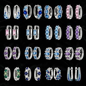 Op de afbeelding: Een verzameling zilveren oorringen met diverse edelsteenversieringen. De oorbellen hebben een verscheidenheid aan kleuren, waaronder blauw, roze, paars en groen. De edelstenen zijn in verschillende vormen en maten gezet, wat een gevarieerde weergave creëert.
