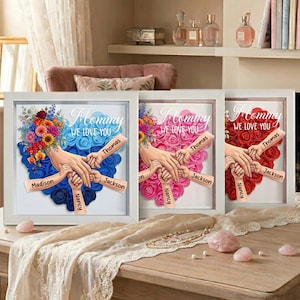 May include: Three framed art pieces with the text "Mommy WE LOVE YOU". Each features a heart-shaped floral arrangement in blue, pink, and red, with hands holding each other. Names are written on the hands. White frames.