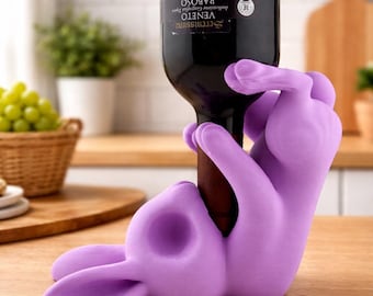 May include: A lavender-colored bunny-shaped wine bottle holder. The bunny is lying on its back, holding a wine bottle. The bottle is dark with a label. The holder is made of a soft, flexible material. The background includes a kitchen setting.