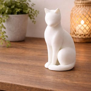 Può includere: Una statuetta di gatto in ceramica bianca in posizione seduta. Il gatto ha una finitura liscia e opaca ed è posto su una superficie di legno. Una pianta in vaso e una lanterna intrecciata sono sullo sfondo.