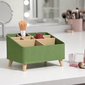 May include: A green makeup organizer with multiple compartments and wooden legs. A makeup brush and lipstick are inside the organizer. Other makeup items, including a brush, lipstick, and lip liner, are on the white surface.