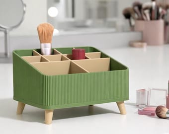 May include: A green makeup organizer with multiple compartments and wooden legs. A makeup brush and lipstick are inside the organizer. Other makeup items, including a brush, lipstick, and lip liner, are on the white surface.