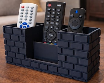 May include: A navy blue remote control organizer with a brick-like design, holding three remotes. The organizer is made of a dark material and sits on a wooden surface. The remotes are black and white.