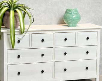 May include: A white wooden dresser with eight drawers and black knobs. A potted plant with green and white striped leaves and a light green owl figurine sit on top of the dresser.