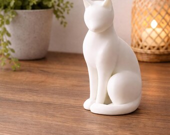 May include: A white ceramic cat figurine in a seated position. The cat has a smooth, matte finish and is placed on a wooden surface. A potted plant and a woven lantern are in the background.