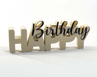 May include: A cream-colored, freestanding "HAPPY" sign with a black cursive "Birthday" overlay. The letters are thick and blocky, creating a 3D effect. The sign is designed for birthday celebrations and is set against a white background.