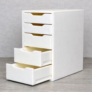 May include: A white, five-drawer storage unit with a rectangular shape. Two drawers are open, revealing a light interior. The unit is made of a solid, opaque material and sits on a gray surface.