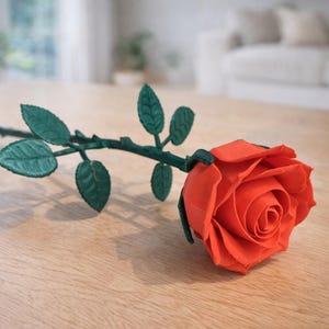 May include: A single, vibrant orange rose with green leaves and stem lies on a light-colored wooden surface. The rose is in full bloom, showcasing detailed petals. The stem and leaves are a deep green, creating a striking contrast.