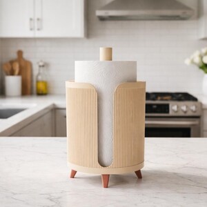 Può includere: Un portarotolo di carta assorbente beige chiaro con un design a coste verticali. Il supporto ha un tassello di legno sulla parte superiore e poggia su tre piccole gambe marrone rossiccio. L'ambiente cucina comprende un piano di lavoro e armadi bianchi.