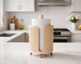 May include: A light beige paper towel holder with a vertical ribbed design. The holder has a wooden dowel on top and rests on three small, reddish-brown legs. The kitchen setting includes a white countertop and cabinets.