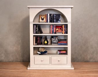 May include: A white miniature bookshelf with an arched top, filled with small books of various colors and sizes. The bookshelf has two drawers at the bottom and is set against a neutral-toned wall.