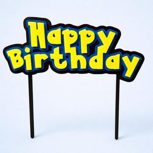 May include: A yellow and blue "Happy Birthday" cake topper with a black border and two black sticks. The text is in a bold, playful font, perfect for a birthday celebration.