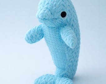 May include: A light blue knitted dolphin plush toy with a textured surface. The dolphin has a black eye and outstretched arms, and a tail fin. The toy is standing upright against a light blue background.
