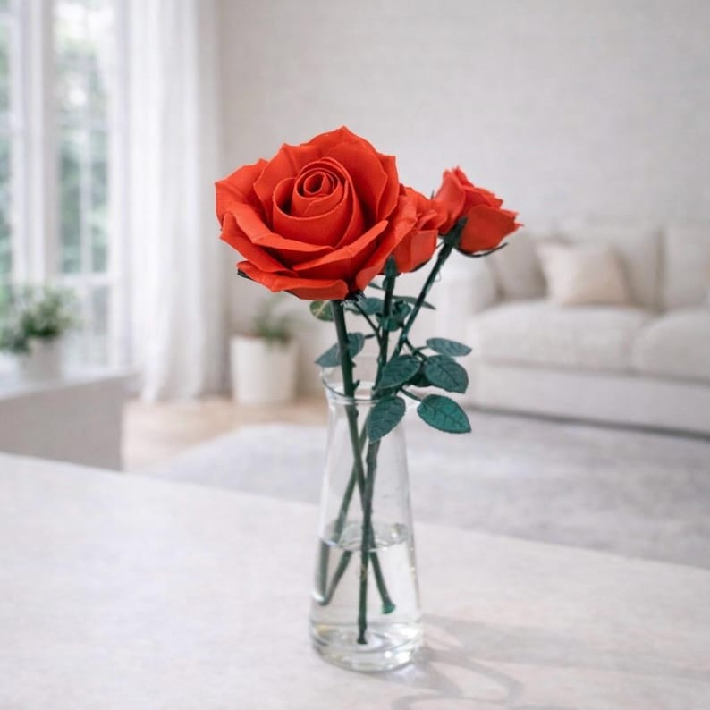 May include: A clear glass vase holds three red roses with green stems and leaves. The roses are in full bloom, with a large central flower and two smaller buds. The vase sits on a white surface, with a blurred background of a window and a couch.