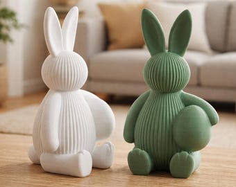 May include: Two decorative ceramic bunny figurines. One is white, the other is green and holding an egg. Both have a ribbed texture and are sitting on a wooden surface. The bunnies are a charming addition to Easter decor.