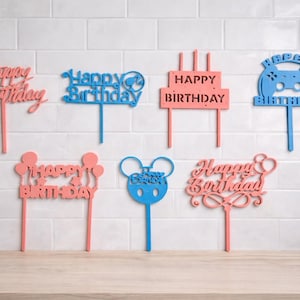 Op de afbeelding: Een verzameling lasergesneden verjaardagstaarttoppers in verschillende ontwerpen en kleuren. De toppers bevatten de zinnen "Happy Birthday" en een vorm van een videogamecontroller. Kleuren zijn onder meer roze en blauw.
