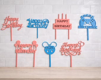 May include: A collection of laser-cut birthday cake toppers in various designs and colors. The toppers include the phrases "Happy Birthday" and a video game controller shape. Colors include pink and blue.