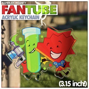 Op de afbeelding: Acryl sleutelhanger met twee stripfiguren: een groene reageerbuis met een straalpistool en een rode waaier met een vlinderdas. De sleutelhanger is gemarkeerd met "FAN TUBE" en meet 8 cm.
