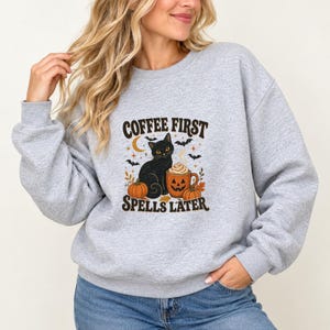 May include: A light gray crewneck sweatshirt featuring a graphic print of a black cat sitting next to a jack-o'-lantern mug and two pumpkins. The text COFFEE FIRST SPELLS LATER is printed in brown above and below the central illustration.