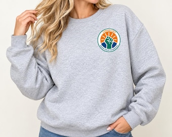 May include: A light gray crewneck sweatshirt featuring a circular logo on the left chest. The logo contains an orange sun graphic, a blue raised fist, and the text 5 MONATE LAUT Bündnis Zukunft Therapieberufe in a circular border.
