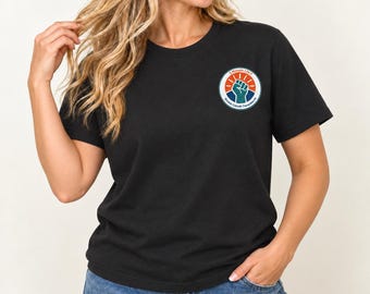 May include: A black short-sleeved t-shirt featuring a circular graphic on the left chest. The design displays a raised fist in teal against a dark blue background, topped with an orange sunburst and the text 5 MONATE LAUT Bündnis Zukunft Therapieberufe.
