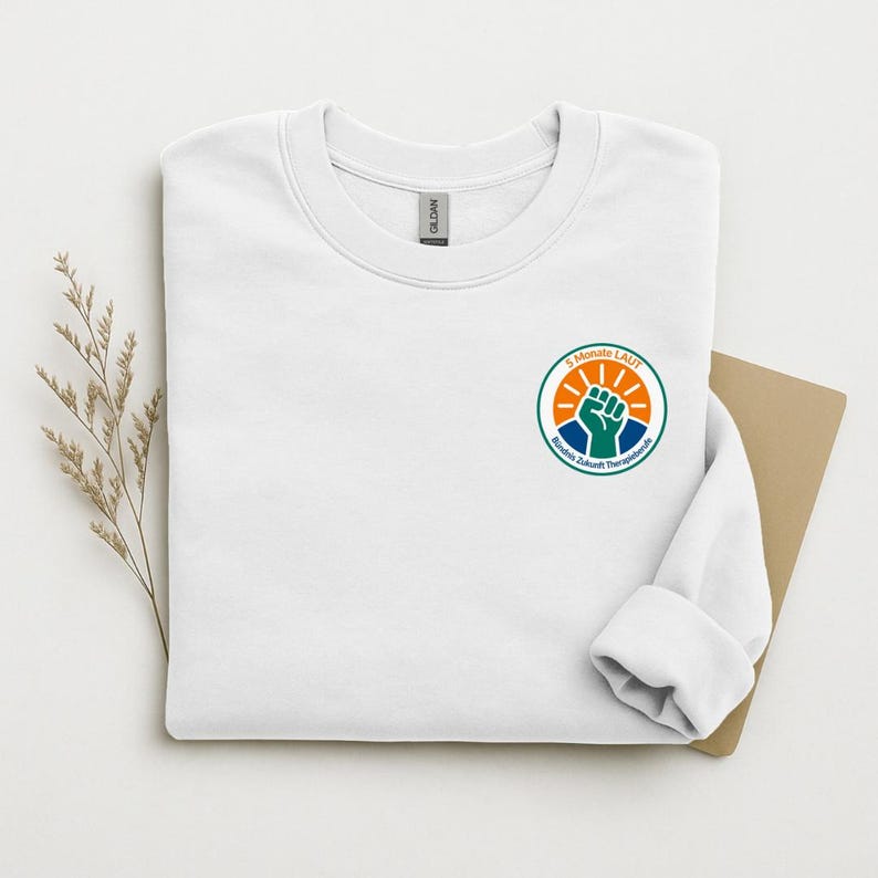 May include: A white crewneck sweatshirt featuring a circular logo on the left chest. The logo displays a green raised fist, an orange sunburst, and blue accents with the text 5 Monate LAUT B&uuml;ndnis Zukunft Therapieberufe printed in black.