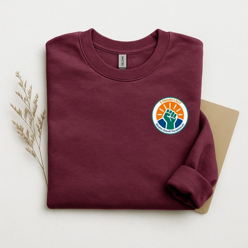 May include: A maroon crewneck sweatshirt featuring a circular graphic on the left chest. The graphic displays a raised fist in green, orange sun rays, and the text 5 MONATE LAUT B&uuml;ndnis Zukunft Therapieberufe in a white and green border.