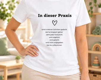 May include: A white short-sleeved t-shirt featuring black text that reads In dieser Praxis, followed by a heart icon and a list of German phrases including wird in kleinen Schritten gedacht, darf es langsam gehen, zählt jeder Fortschritt, wird zugehört, wird gelacht, wird nicht aufgegeben, bist du willkommen.