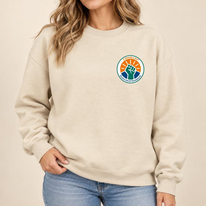 May include: A beige crewneck sweatshirt featuring a circular logo on the left chest. The logo contains an orange sun graphic, a teal raised fist, and the text 5 MONATE LAUT B&uuml;ndnis Zukunft Therapieberufe in a circular blue border.