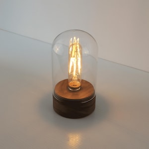 May include: A decorative table lamp featuring a clear glass dome over a warm, glowing Edison-style light bulb. The bulb sits on a round, wooden base with a dark brown finish. The lamp is designed for ambient lighting.