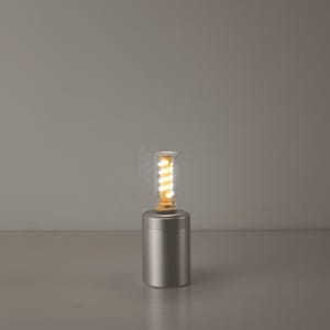 Op de afbeelding: Een moderne lamp met een zilverkleurige cilindrische voet en een heldere glazen lamp. De lamp bevat een warm, spiraalvormig LED-licht. De lamp is ontworpen voor een minimalistische stijl en biedt zachte omgevingsverlichting.
