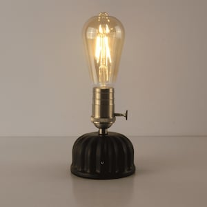 May include: A vintage-style table lamp featuring a large, Edison-style light bulb. The bulb is clear with visible filaments, emitting a warm, golden glow. The lamp has a metallic base and a black, ribbed bottom.