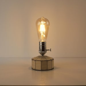 Könnte beinhalten: Eine Tischlampe im Vintage-Stil mit einer großen, klaren Edison-Glühbirne, die ein warmes Leuchten ausstrahlt. Die Lampe hat einen silbernen Metallsockel und einen geometrischen Holzschirm. Das Licht ist an.