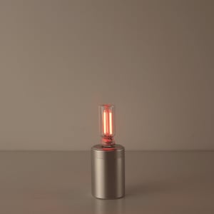 May include: A small, cylindrical, brushed metal lamp base with a clear glass bulb. The bulb contains two vertical, glowing red filaments. The lamp is designed to sit on a flat surface and emits a warm, red light.