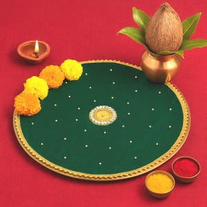 May include: A circular dark green decorative tray featuring a gold scalloped border, small white pearl-like accents, and a central circular embellishment with clear crystals. It is accompanied by yellow and orange marigold flowers, a lit clay lamp, a copper pot with a coconut, and two small bowls containing red and yellow powders.