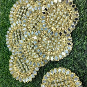 Può includere: Una applicazione ricamata color oro caratterizzata da motivi circolari decorati con piccoli specchietti riflettenti e minuscole perline bianche. Il design include un pezzo più grande a più cerchi e una piccola toppa circolare separata.