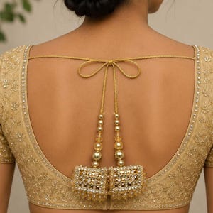 Op de afbeelding: Goudkleurige blouse met korte mouwen en ingewikkelde borduursels en versieringen. De achterkant heeft een striksluiting en hangende kralenkwasten. De blouse is versierd met kleine, glinsterende details, wat een luxueuze en elegante uitstraling creëert.