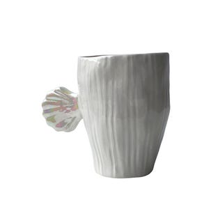 Puede incluir: Una taza de cerámica blanca con una superficie texturizada y un asa en forma de concha. El asa tiene detalles iridiscentes. La taza tiene forma cilíndrica y está sobre un fondo blanco.