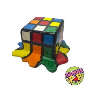 Puede incluir: Una colorida escultura de un Cubo de Rubik derritiéndose. Los cuadrados del cubo están pintados en varios colores, con la pintura goteando por los lados. La escultura tiene un contorno negro y se encuentra sobre un fondo blanco. Un logotipo está en la esquina inferior derecha.