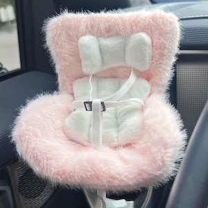May include: A plush pink car seat cover with a white headrest and seat padding. The seat has a white harness with a black buckle. The seat cover is designed to fit in a car.
