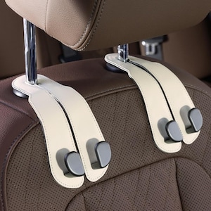 May include: Two cream-colored car seat headrest hooks with gray hooks attached to a brown leather car seat. The hooks are designed to hang items in a vehicle. The headrest is tan.