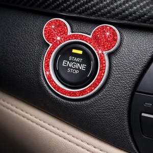 May include: A car's start/stop button is framed by a red, glittery, mouse-ear-shaped bezel. The button is black with white text that reads "START ENGINE STOP" and a yellow indicator light. The button is set in a black leather interior.