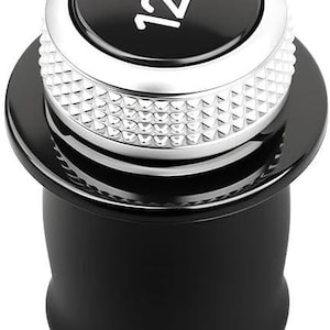 Puede incluir: Una toma de corriente de 12V negra y plateada. La parte superior es negra con texto blanco, rodeada por un anillo plateado y una banda plateada texturizada. La base es un cilindro negro liso y redondeado.